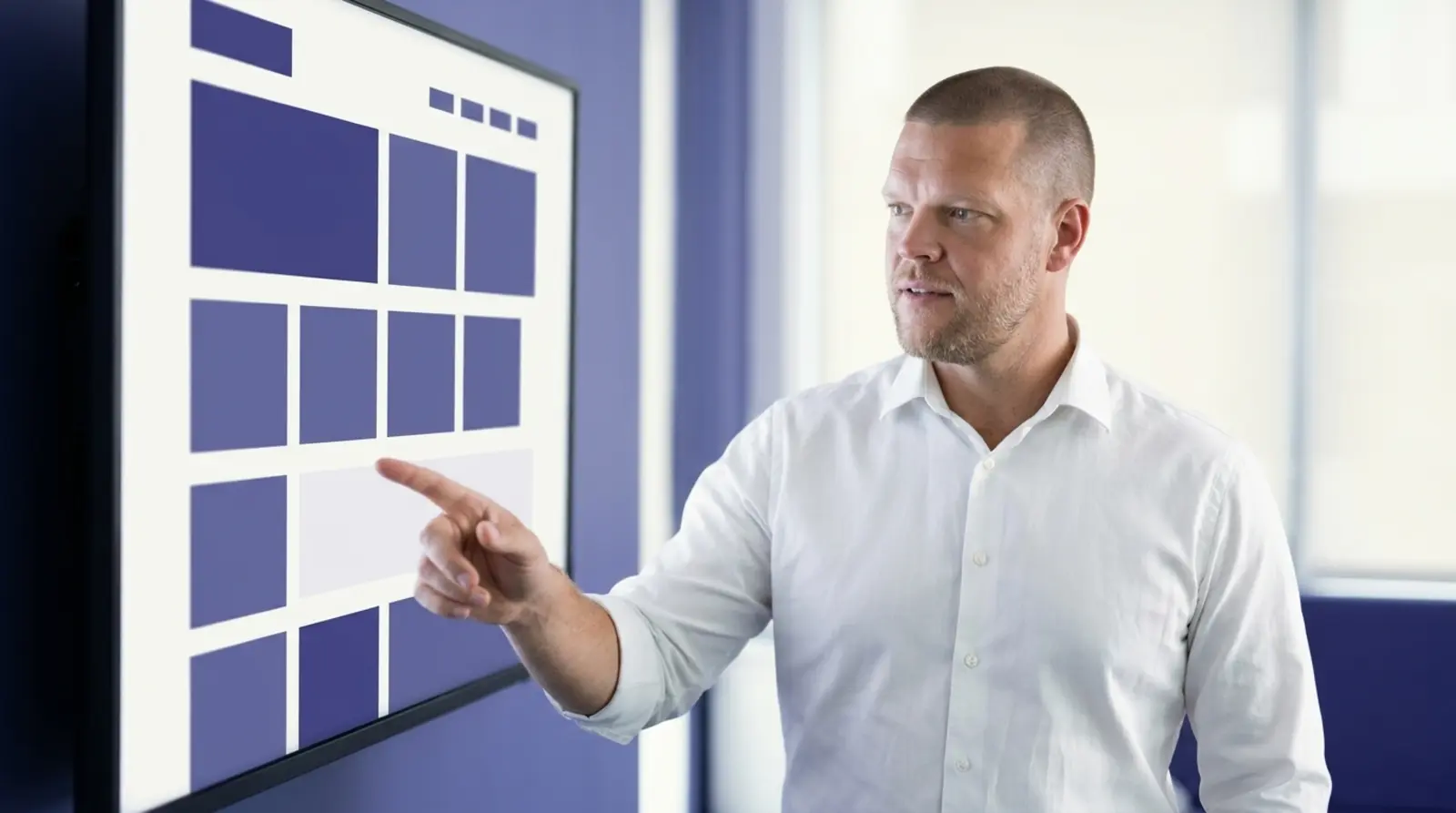 Mike Moore, founder of Strategic AI Architects, in a white shirt pointing at a wall-mounted screen showing an abstract indigo and white website wireframe layout made of simple rectangles, standing in a minimalist indigo-toned office