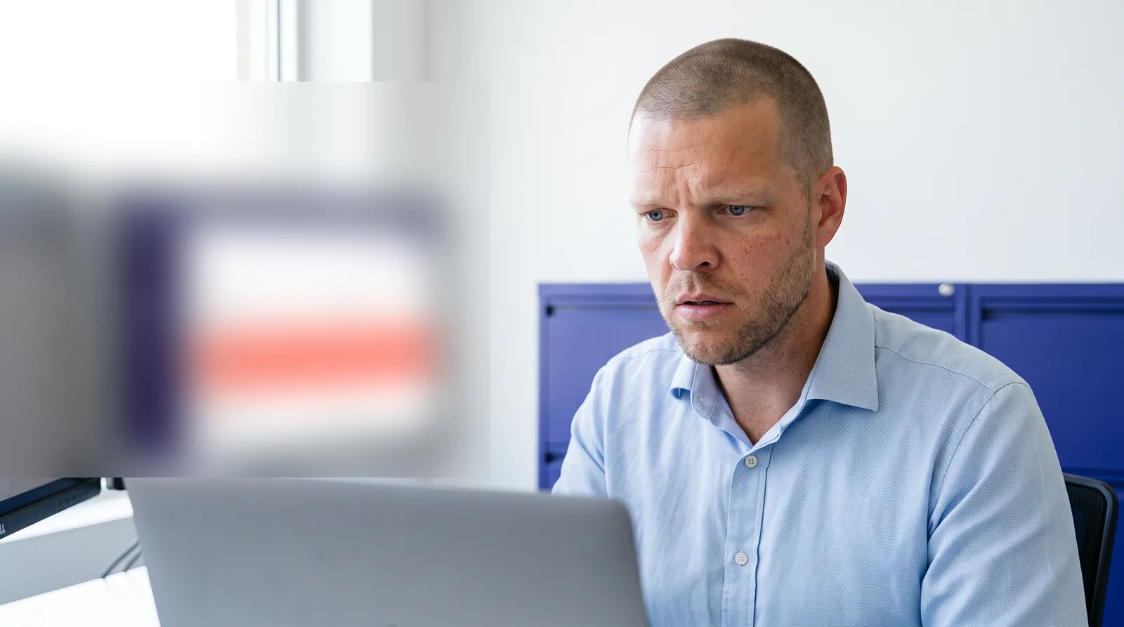 An insurance agency founder with closely buzzed short hair and light stubble sits at a bright office desk, brow furrowed, looking intently at a laptop screen, with an out of focus second monitor showing soft indigo and coral blocks in the background, representing an agency owner discovering that a workflow silently stopped firing for a merged duplicate contact