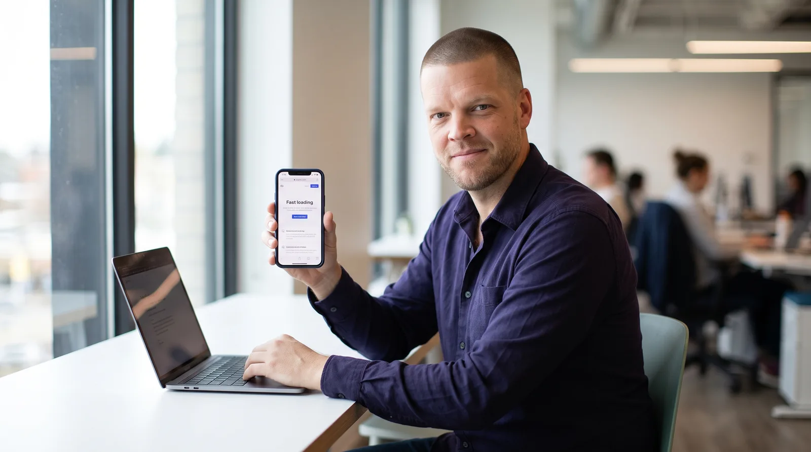 An insurance agency owner in an office holds a smartphone showing a fast loading website while a laptop sits open on the desk beside him, representing the gap between how a site performs on a phone versus a desktop