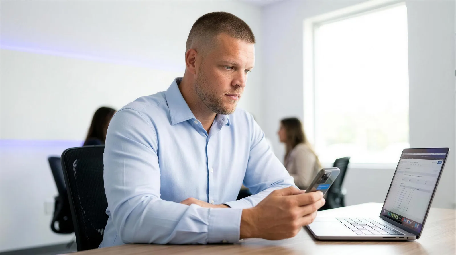 An insurance agency owner with short buzzed hair sits at his desk in a bright office looking at a text message conversation on his phone, with a laptop open beside him showing a CRM lead list, representing the moment an agency owner checks the automated follow up going out to leads